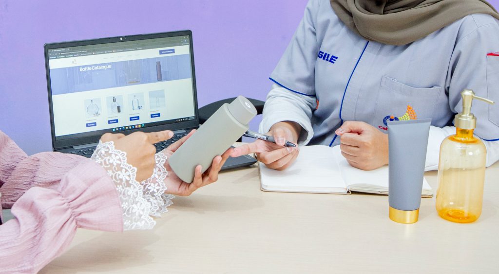 Person in a hijab shows a smartphone to another person while a laptop screen displays a bottle catalogue online.